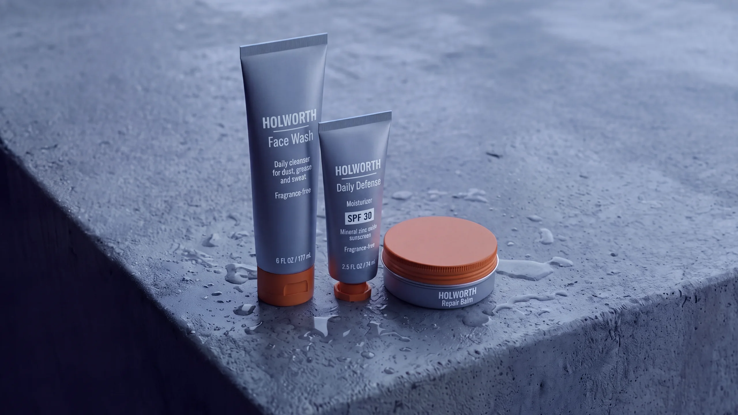 The Holworth Workday Kit on a wet concrete ledge: a tube of Face Wash, a tube of Daily Defense SPF 30 moisturizer and a tin of Repair Balm, steel grey with orange caps.